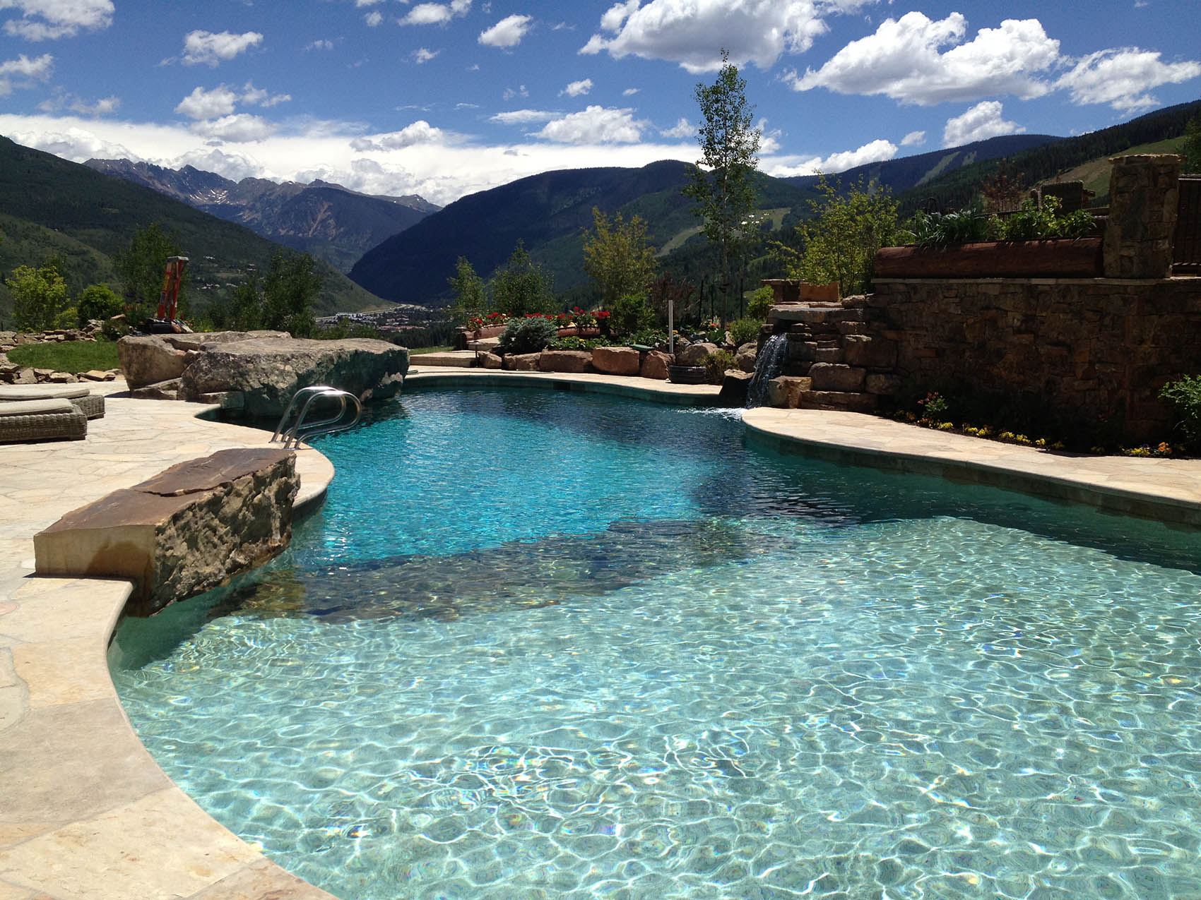 A luxurious outdoor swimming pool with clear blue water, natural stone edges, and a waterfall set against a backdrop of mountains and a clear sky.