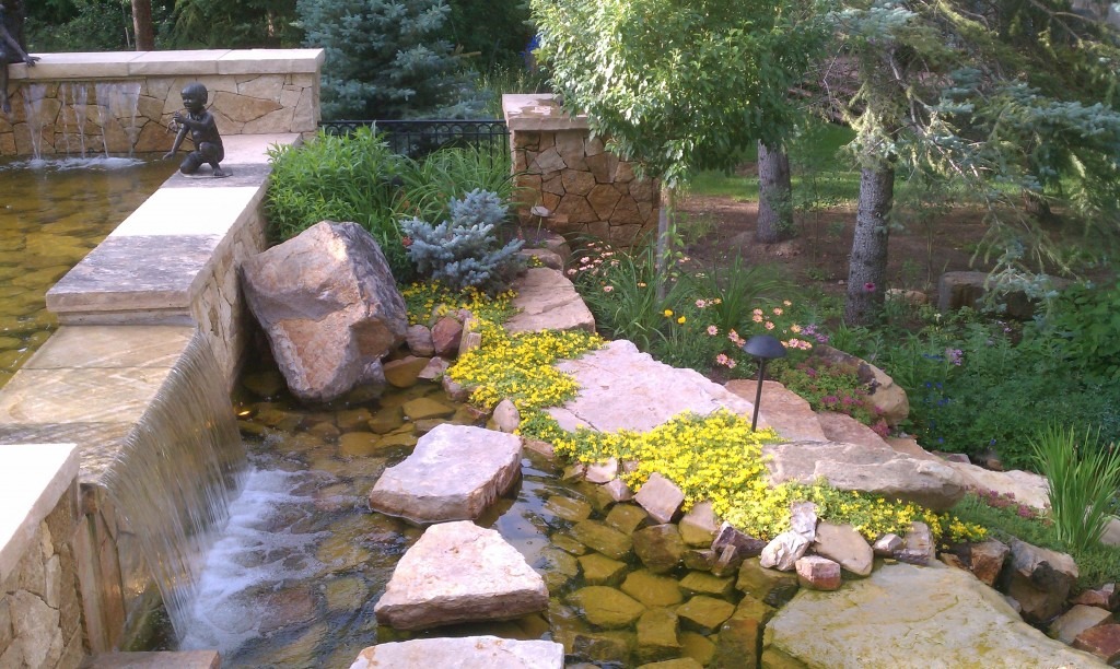 The image shows a tranquil garden setting with a water feature, stone walkways, lush plants, and a sculpture of a child by the water.