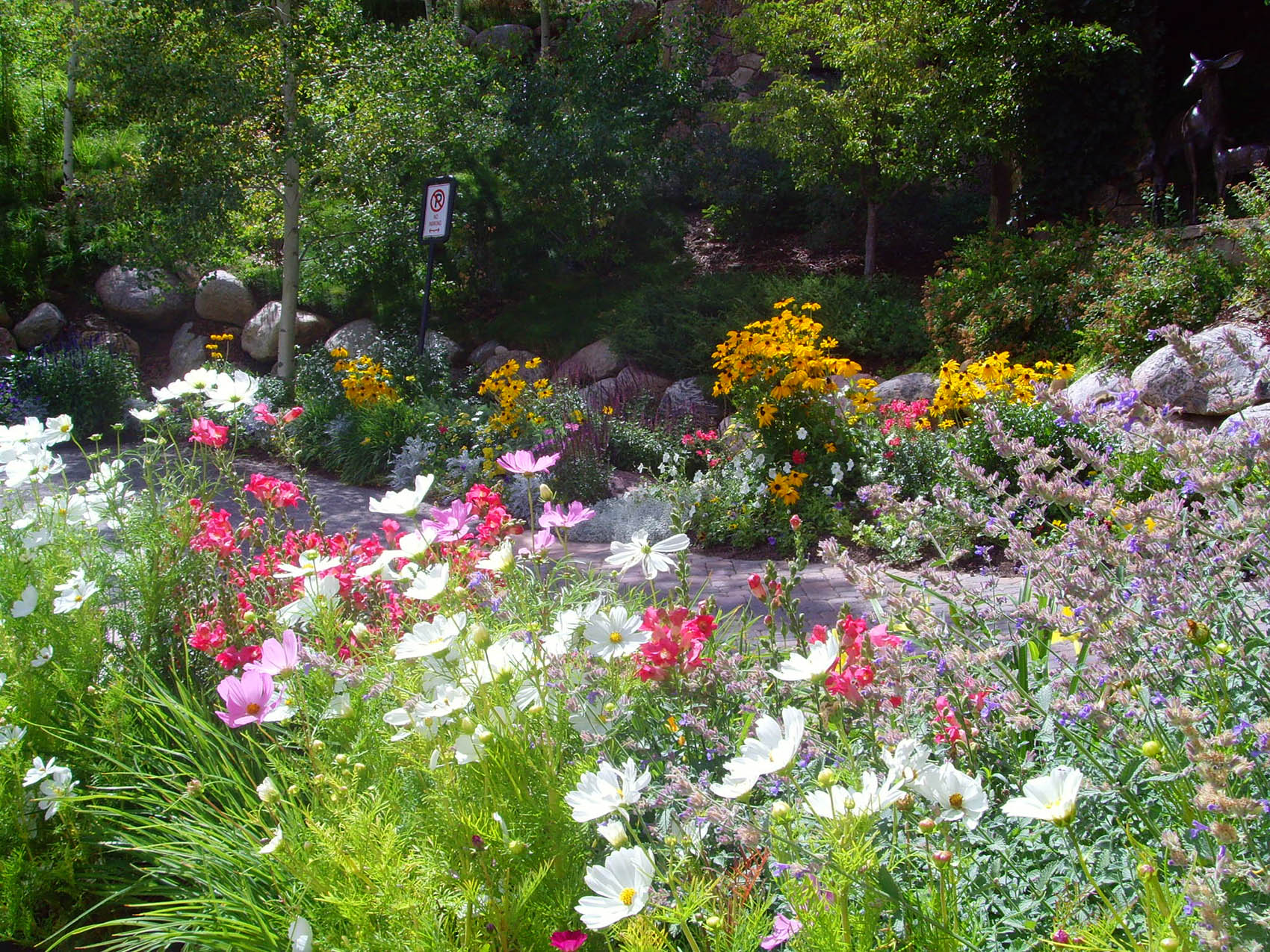 This image showcases a vibrant garden with an abundance of colorful flowers, a footpath, boulders, green foliage, and a statue resembling a deer.
