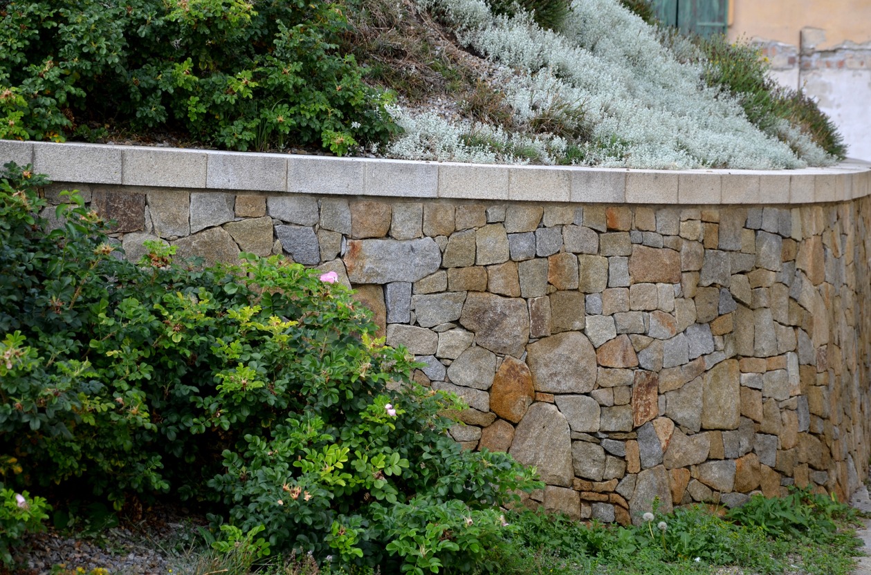 A curved retaining wall made of irregularly shaped stones with a flat capstone. Green shrubs and ground cover plants grow above and around it.