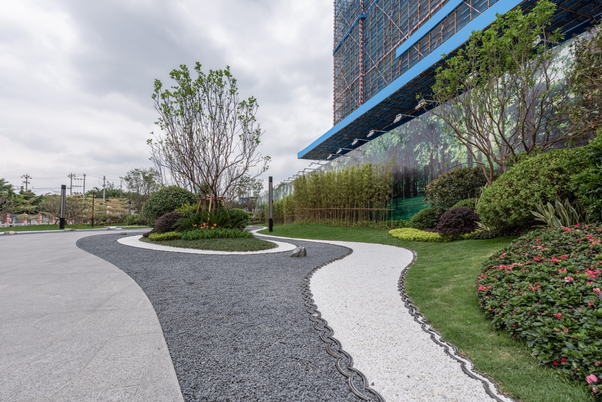 The image shows a modern landscaped garden with winding pathways, green shrubs, and a building adorned with a large forest-themed mural.