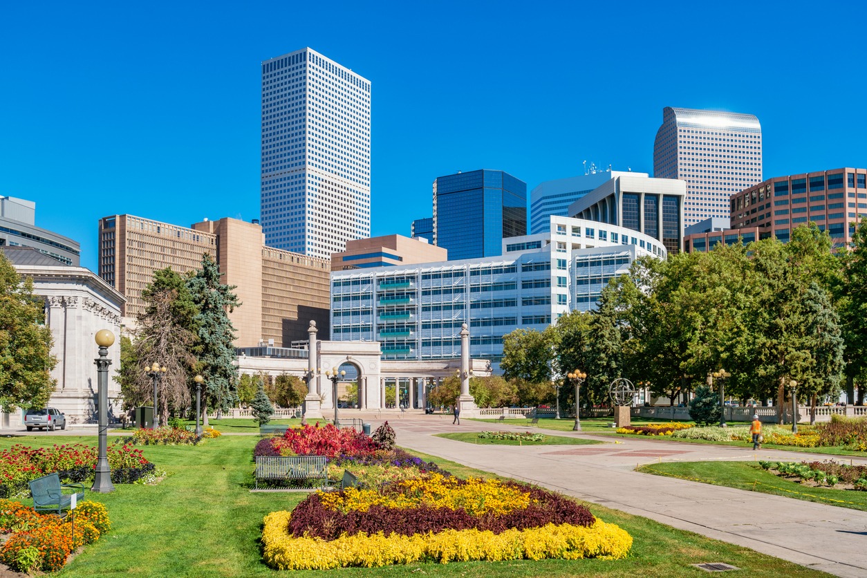 This image shows a sunny urban park with colorful flowerbeds, walking paths, and a backdrop of diverse modern high-rise buildings under a blue sky.