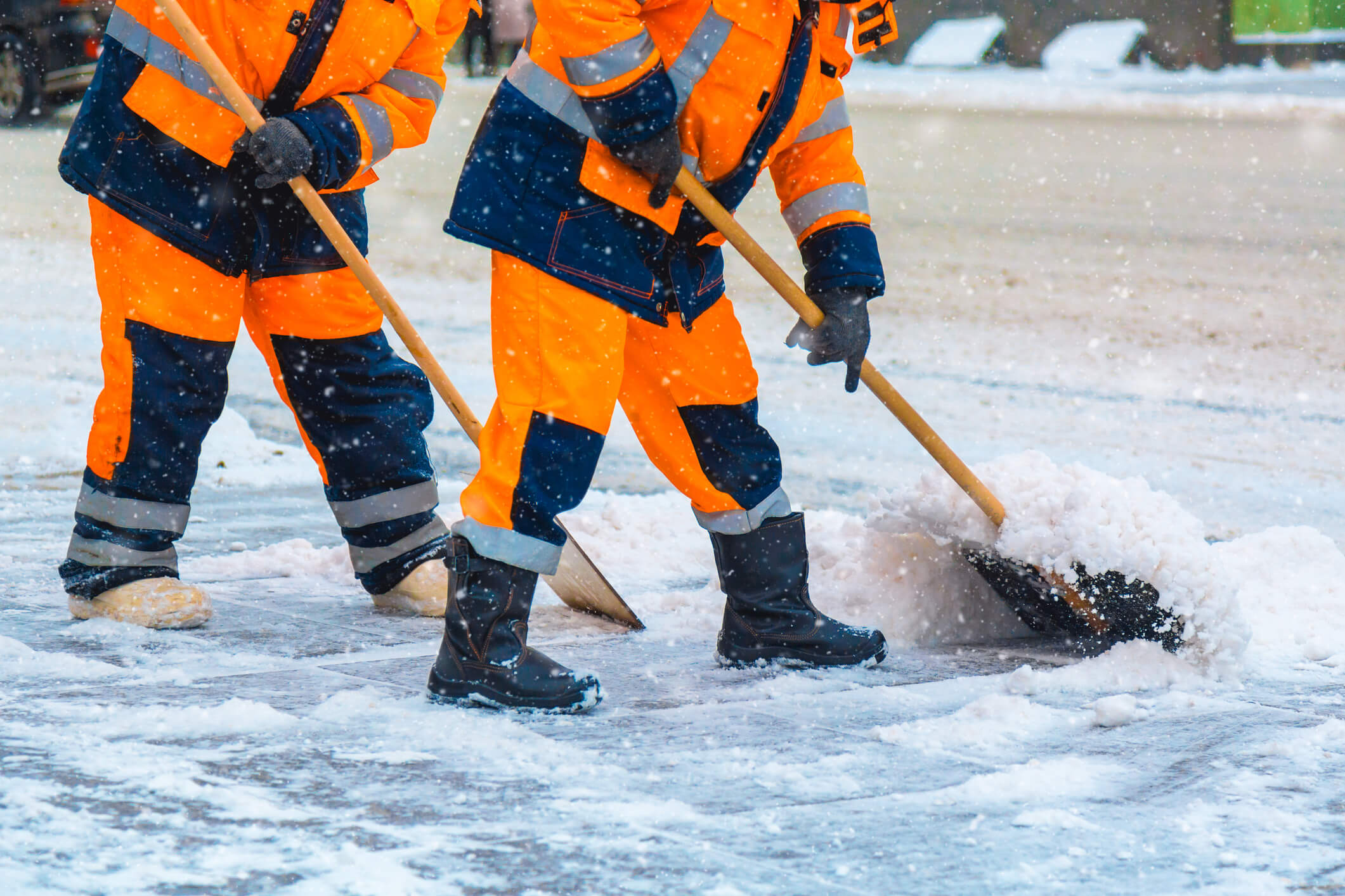 Two workers in orange reflective suits shoveling snow from a paved surface during a snowfall, ensuring clear and safe walkways.